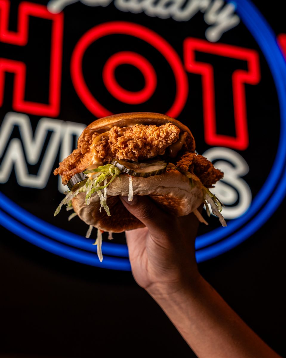 Hand holding a fried chicken sandwich in front of a neon HOT sign